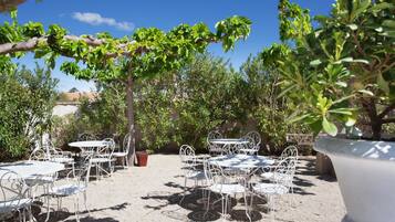 Patio esterno con tavoli e sedie in metallo bianco, immerso nel verde e alberi.
