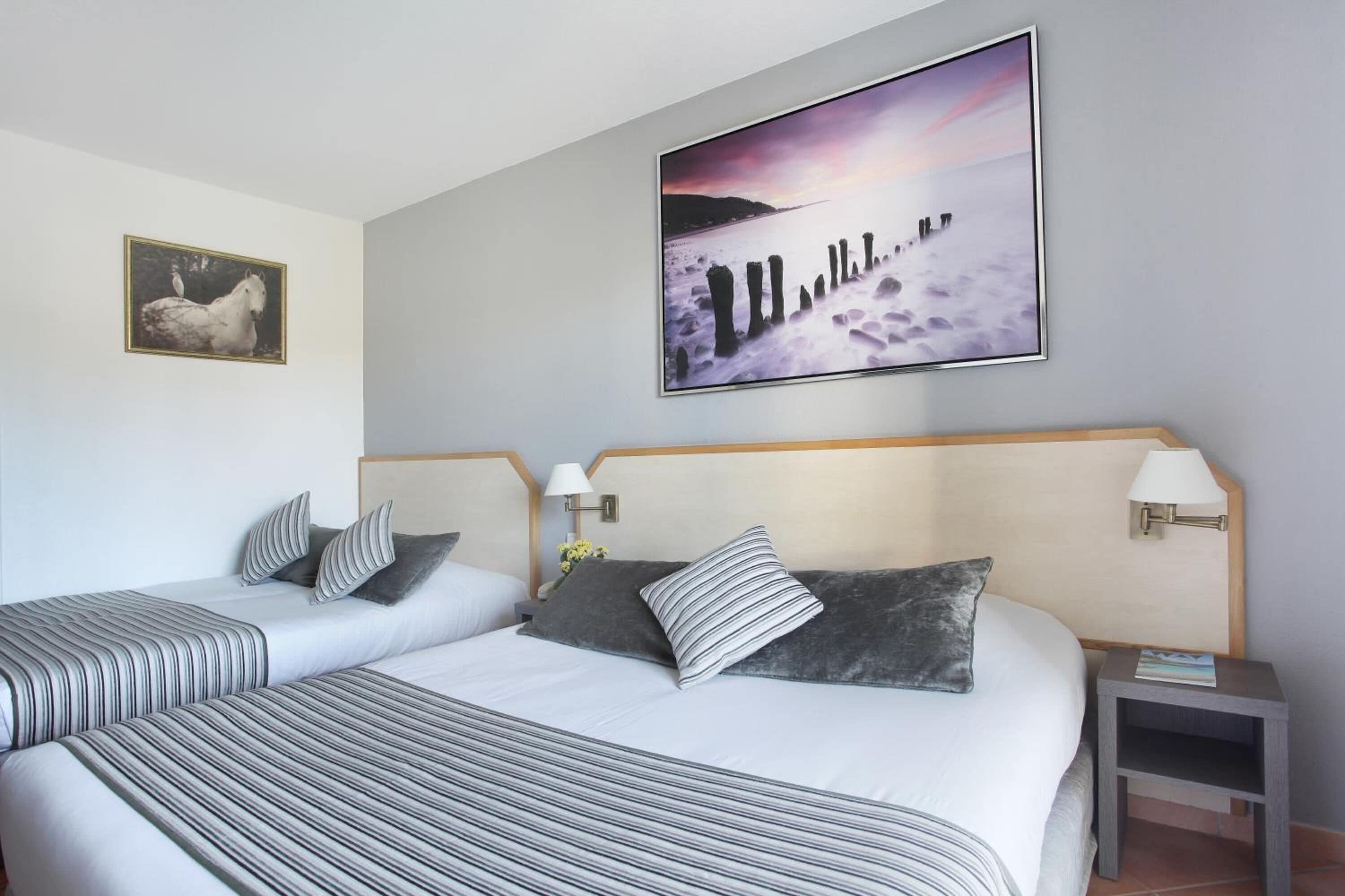 Habitación de hotel con dos camas, un cuadro grande de una escena costera, un retrato enmarcado de un caballo y una pequeña mesa con una lámpara.