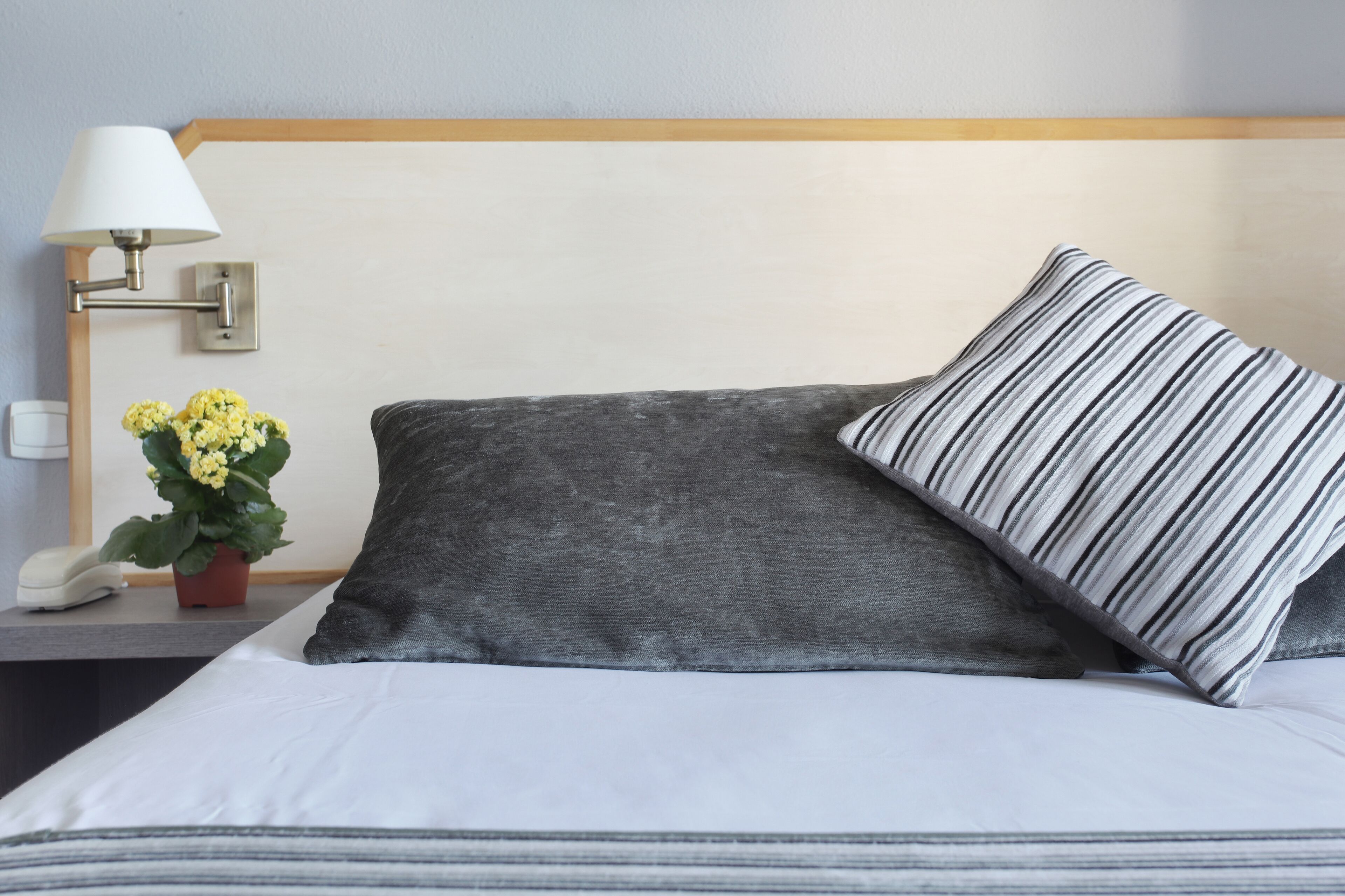 Una cama bien hecha con un almohadón gris y uno a rayas, una lámpara de noche y una pequeña maceta con flores amarillas.