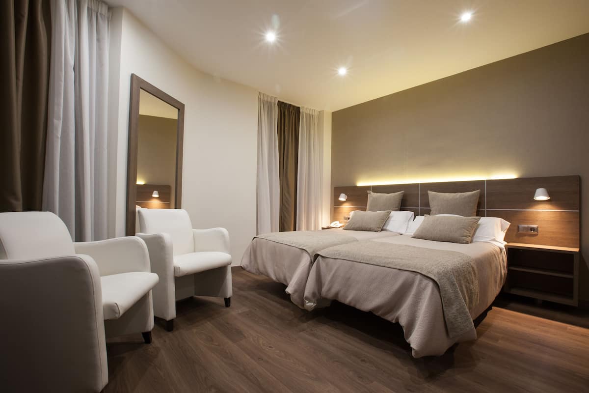 Una habitación de hotel moderna con una cama grande, dos sillones blancos, piso de madera y un espejo grande.
