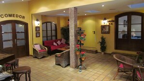 Un interior acogedor con un sofá rojo, sillas de mimbre, suelo de baldosas y un pilar adornado con plantas colgantes.