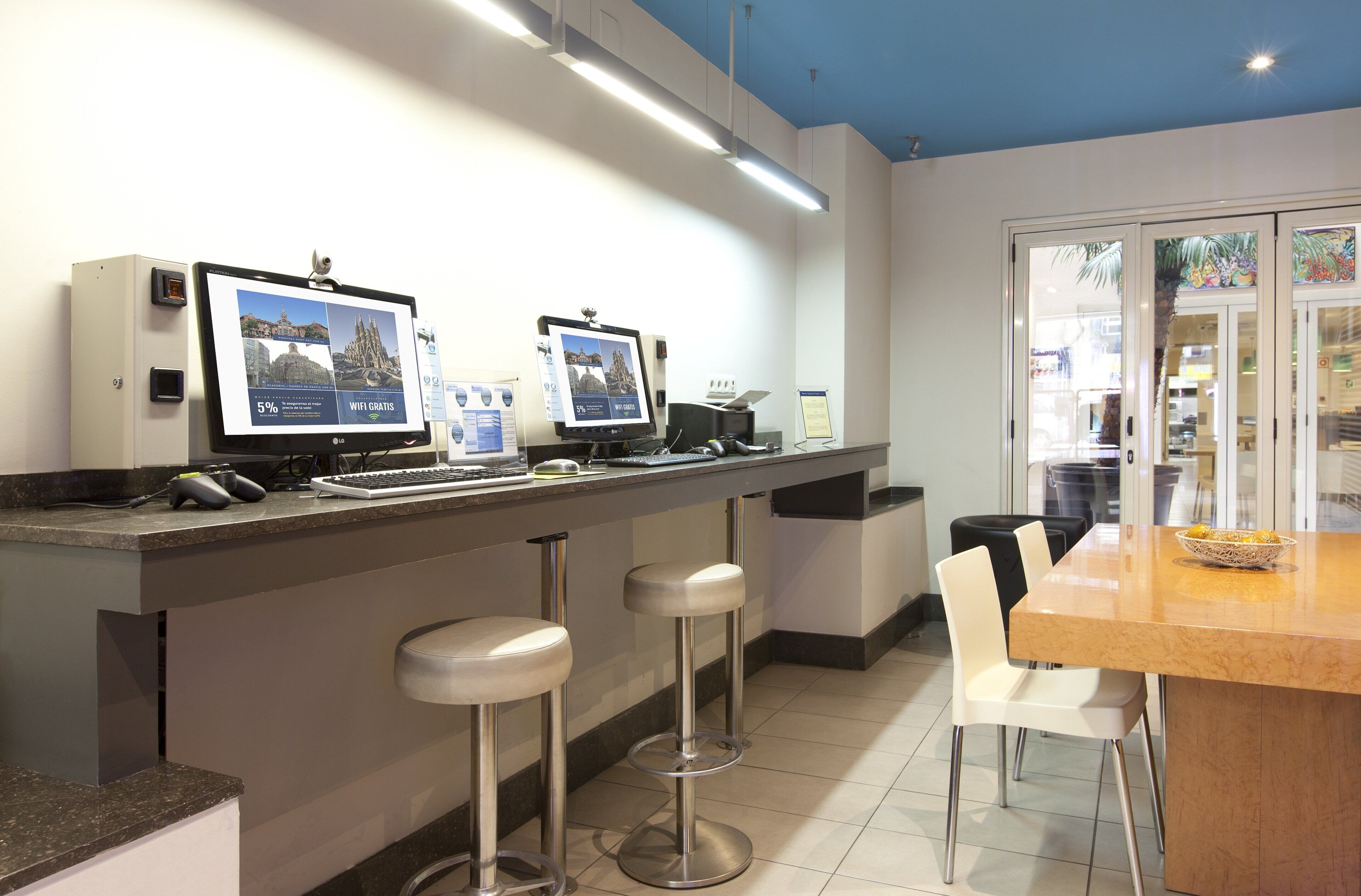 A modern kitchen with two bar stools, a wooden dining table, and two monitors displaying travel information.