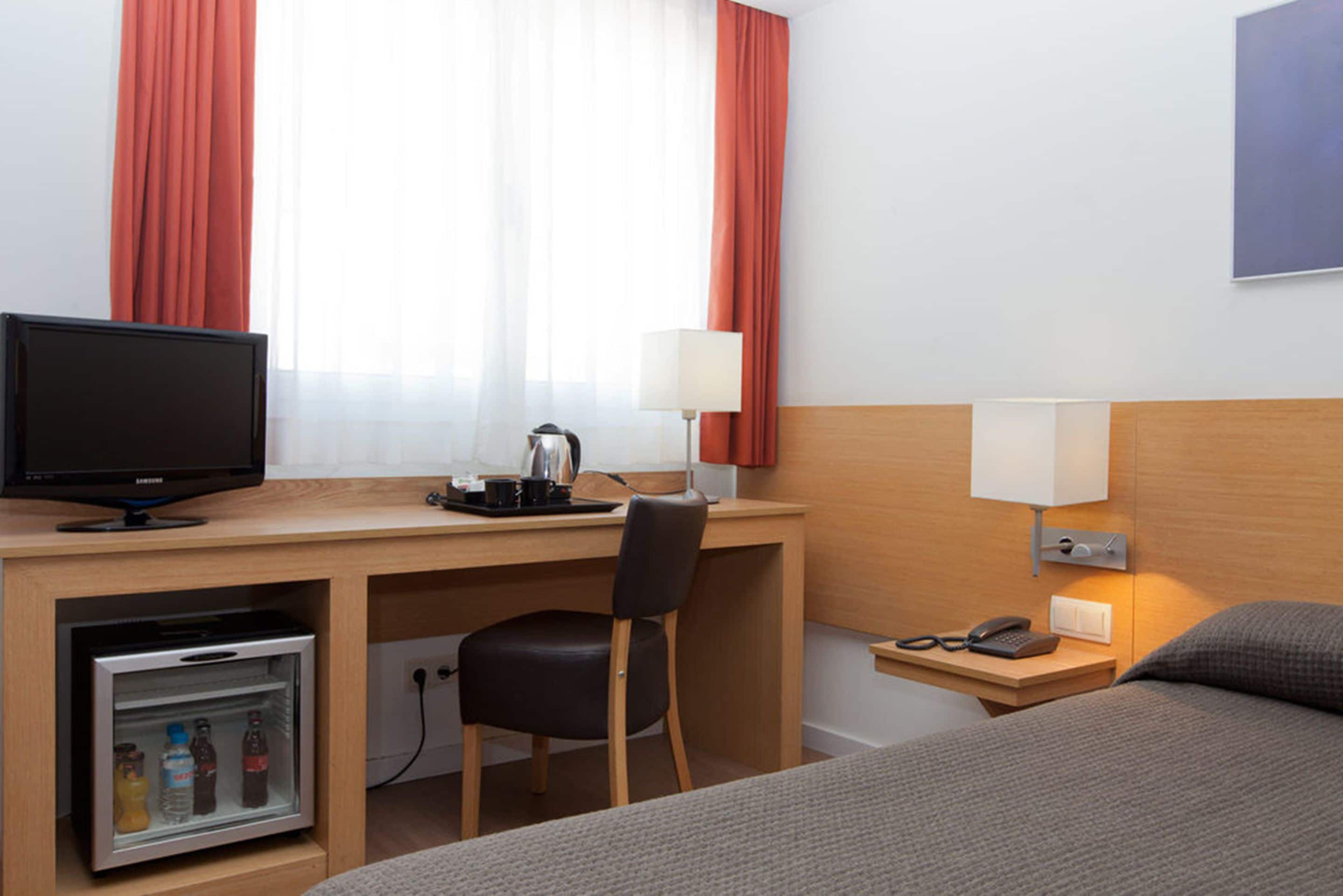 Habitación de hotel con cama, escritorio con computadora, silla, refrigerador pequeño, lámpara y mesita de noche con teléfono y gafas.