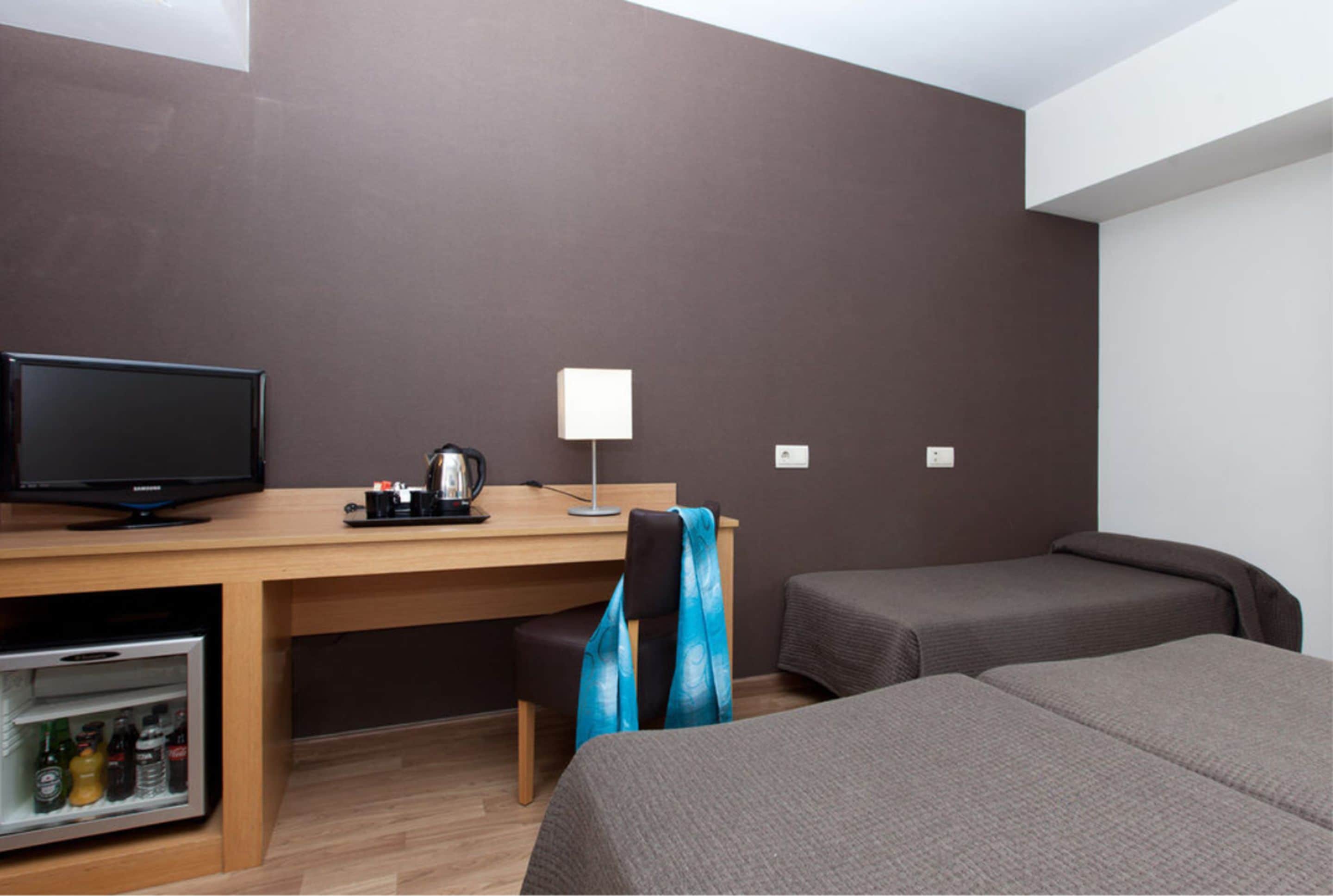 A hotel room with a wooden desk, a television, a lamp, and two single beds.
