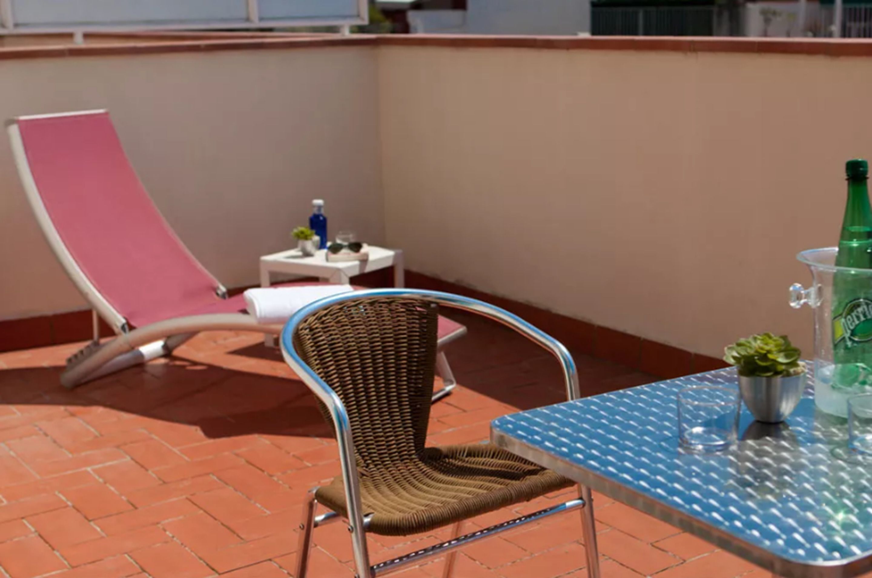Un balcón con una silla de mimbre, una silla lounge rosa, una mesita con una botella y vasos, y una mesa metálica con una planta y una botella.