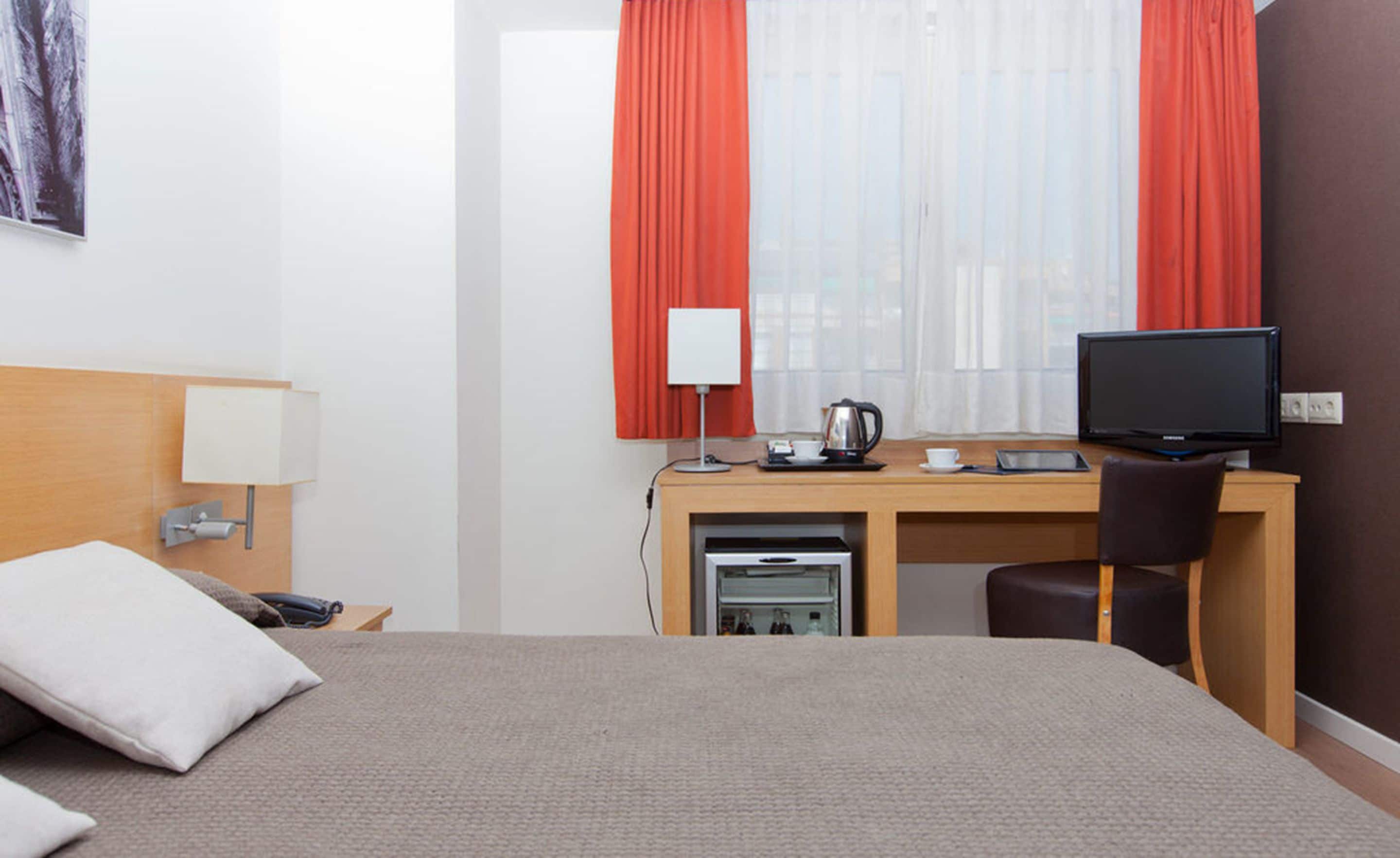 A hotel room with a bed, a desk with a computer, a chair, a lamp, and a window with curtains.