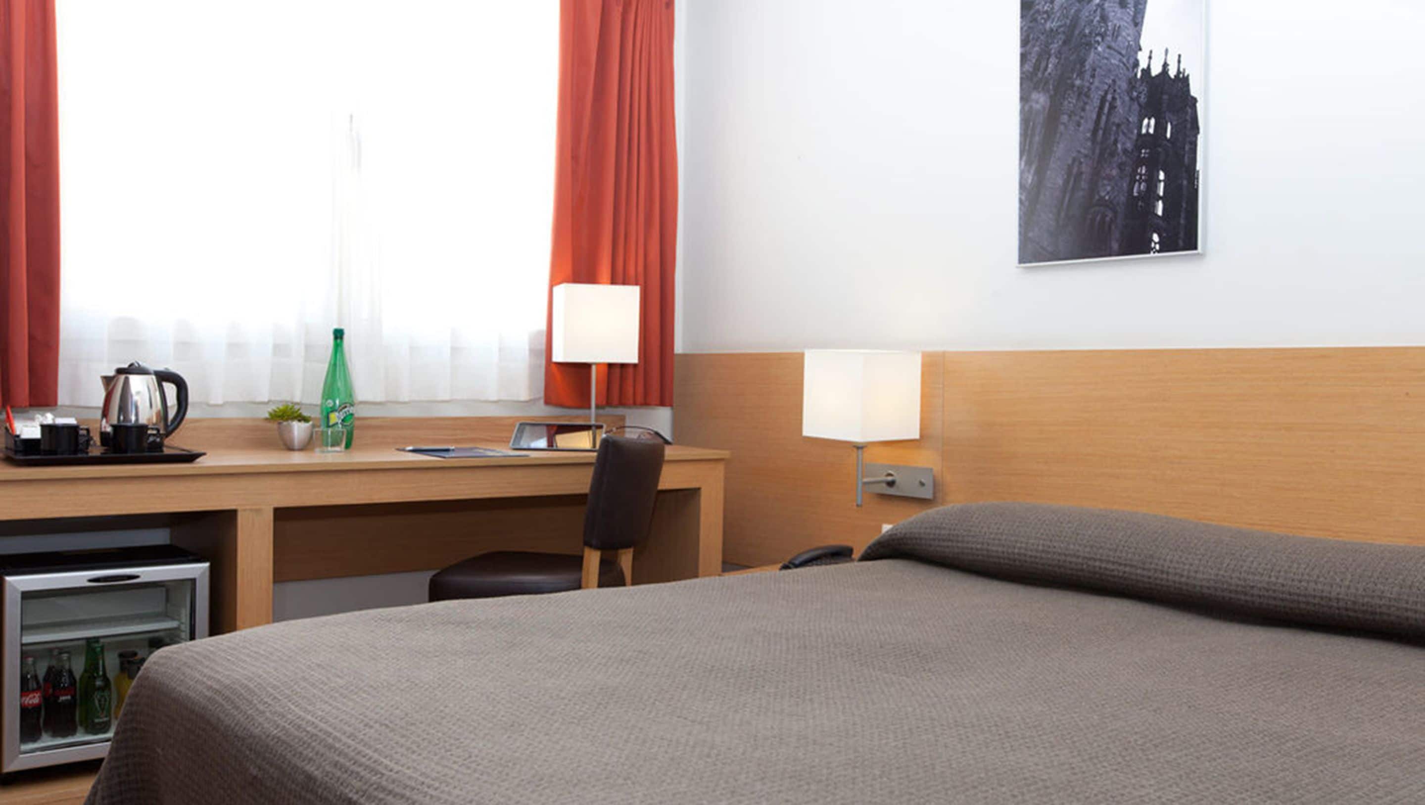 Habitación de hotel con cama, escritorio, silla, lámpara, teléfono y un refrigerador pequeño.