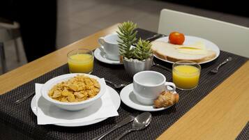 Desayuno con cereal, tostadas, jugo y una pequeña planta sobre una mesa.