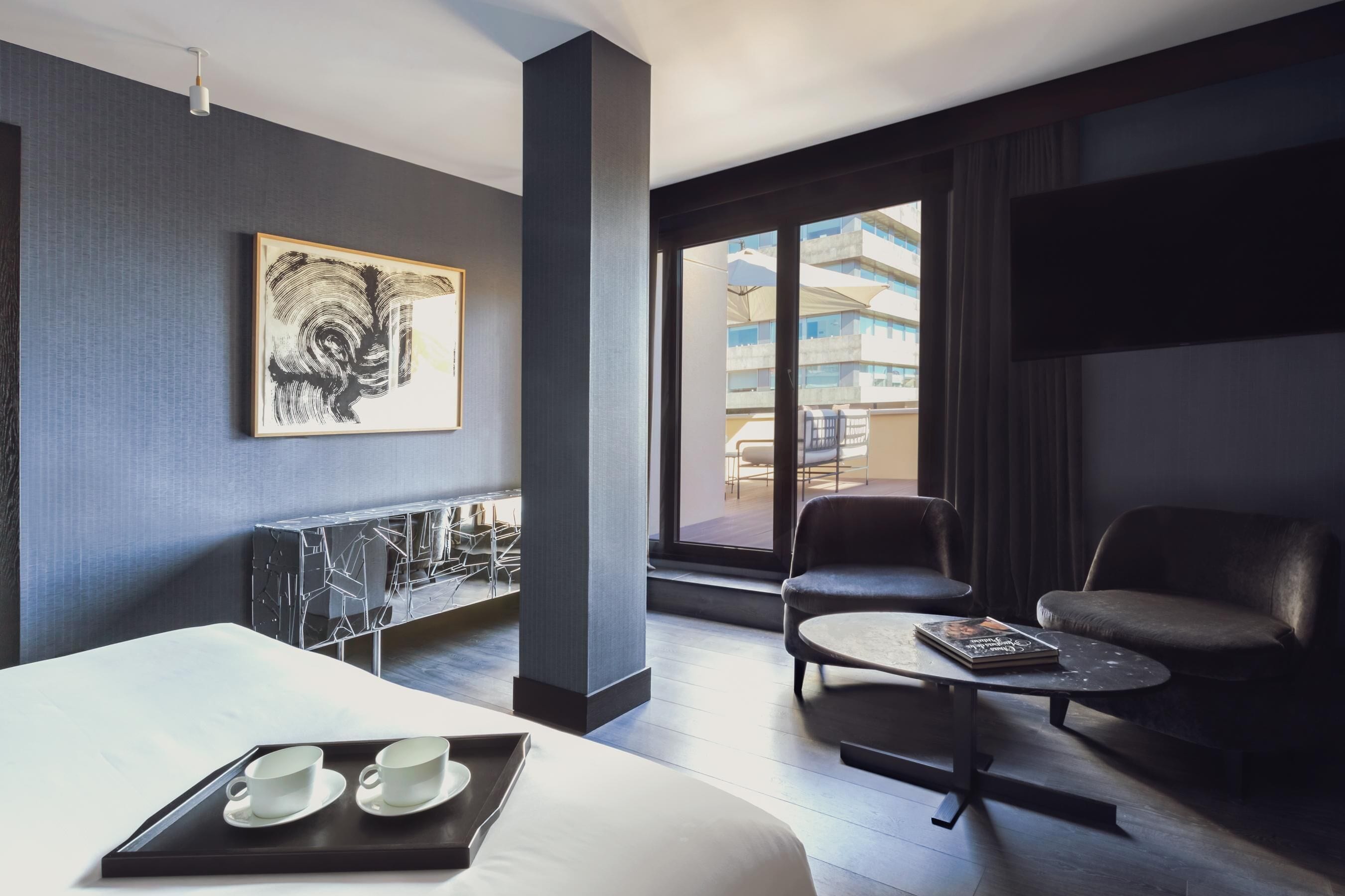 Habitación de hotel moderna con una cama grande, zona de estar con una mesita de café, televisor y un ventanal con vistas a la ciudad.