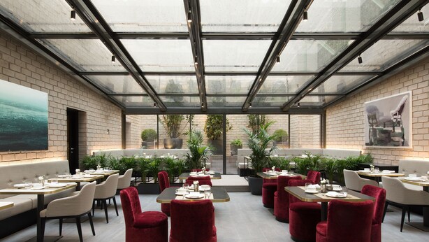 Restaurante moderno com teto de vidro, paredes de tijolo e cadeiras de veludo vermelho. Mesas montadas com pratos e talheres.