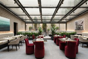 Un restaurante moderno con techo de vidrio, paredes de ladrillo y sillas de terciopelo rojo. Las mesas están dispuestas con platos y cubiertos.