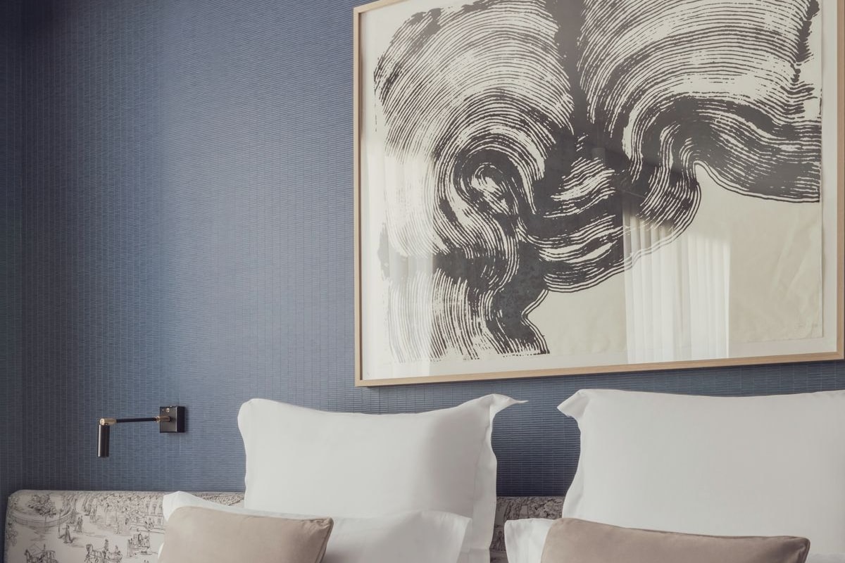 Un dormitorio moderno con una gran obra de arte enmarcada en la pared, una cama con ropa de cama blanca y beige, y una mesita de noche con una lámpara.