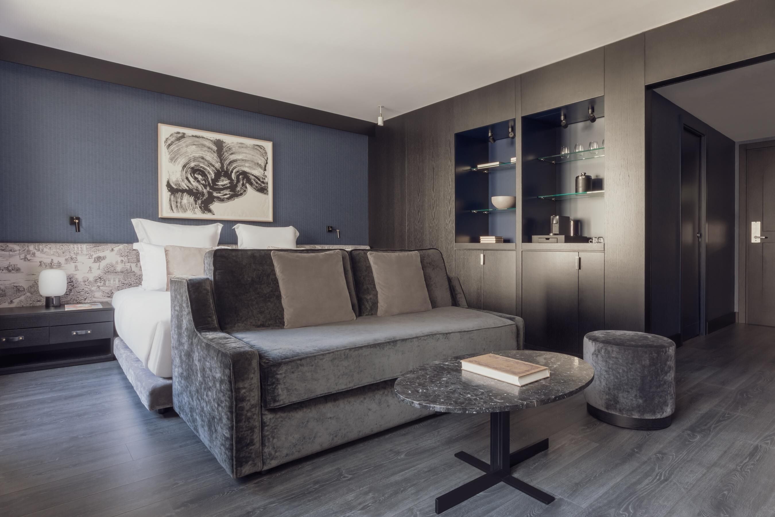 Une chambre moderne avec un canapé gris, une table ronde sur laquelle repose un livre, et un grand tableau abstrait accroché au mur.