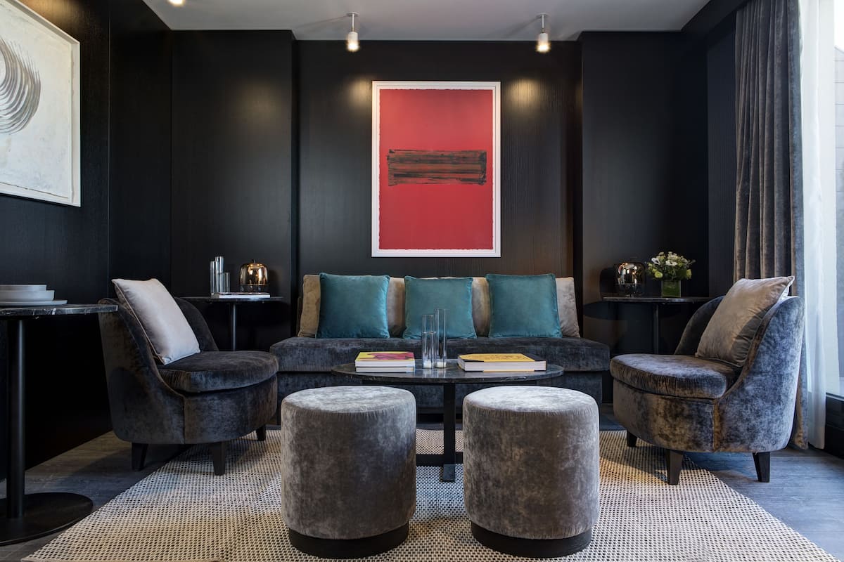 A modern living room with a dark color scheme, featuring a sofa set, two armchairs, and ottomans. A red artwork is displayed on the wall above the sofa.