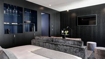Een moderne slaapkamer met een groot bed, inbouwplanken met glazen, een flatscreen televisie en een zithoek met een bank en een voetenbankje.