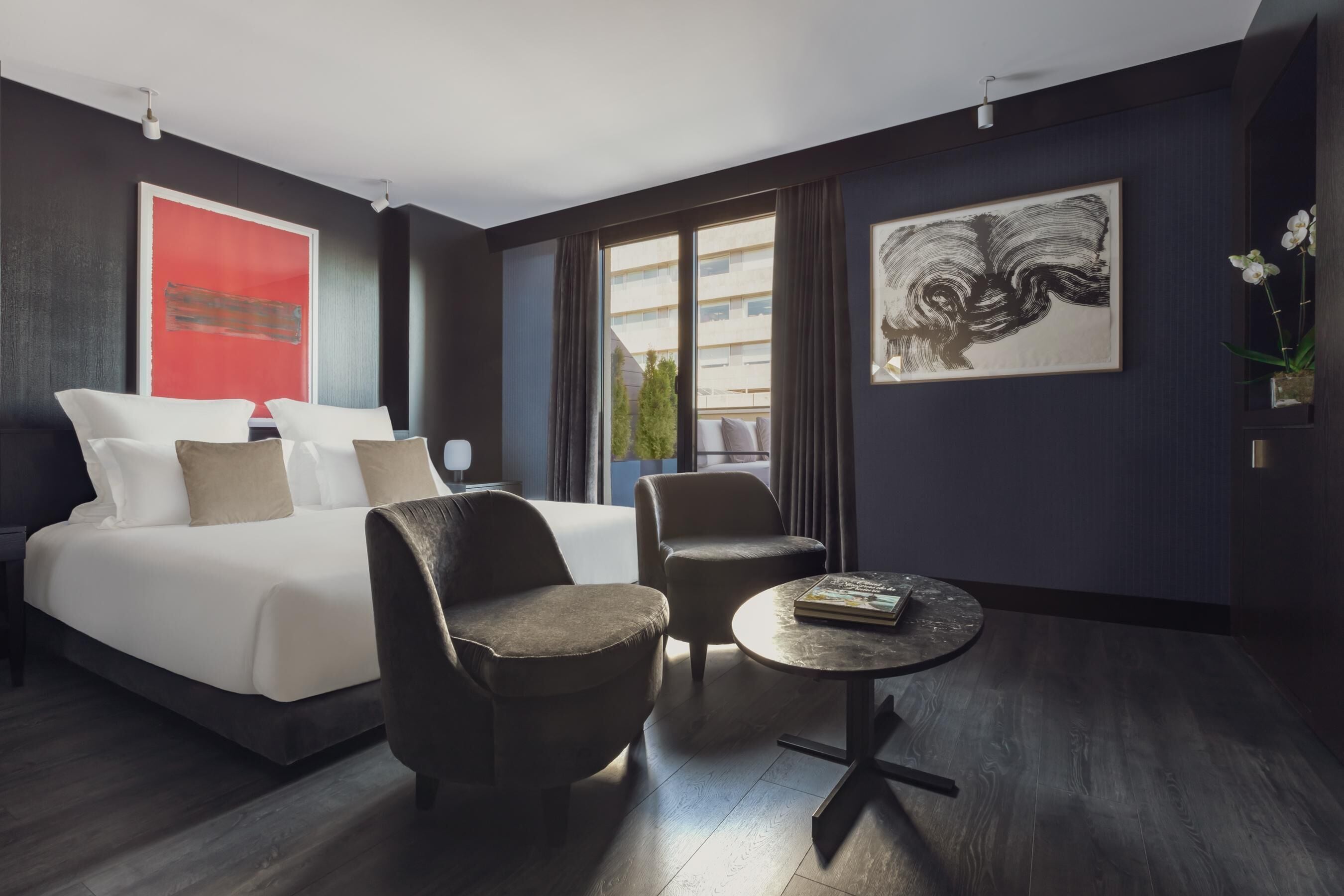 A modern hotel room with a large bed, two armchairs, a small round table, and artwork on the walls.