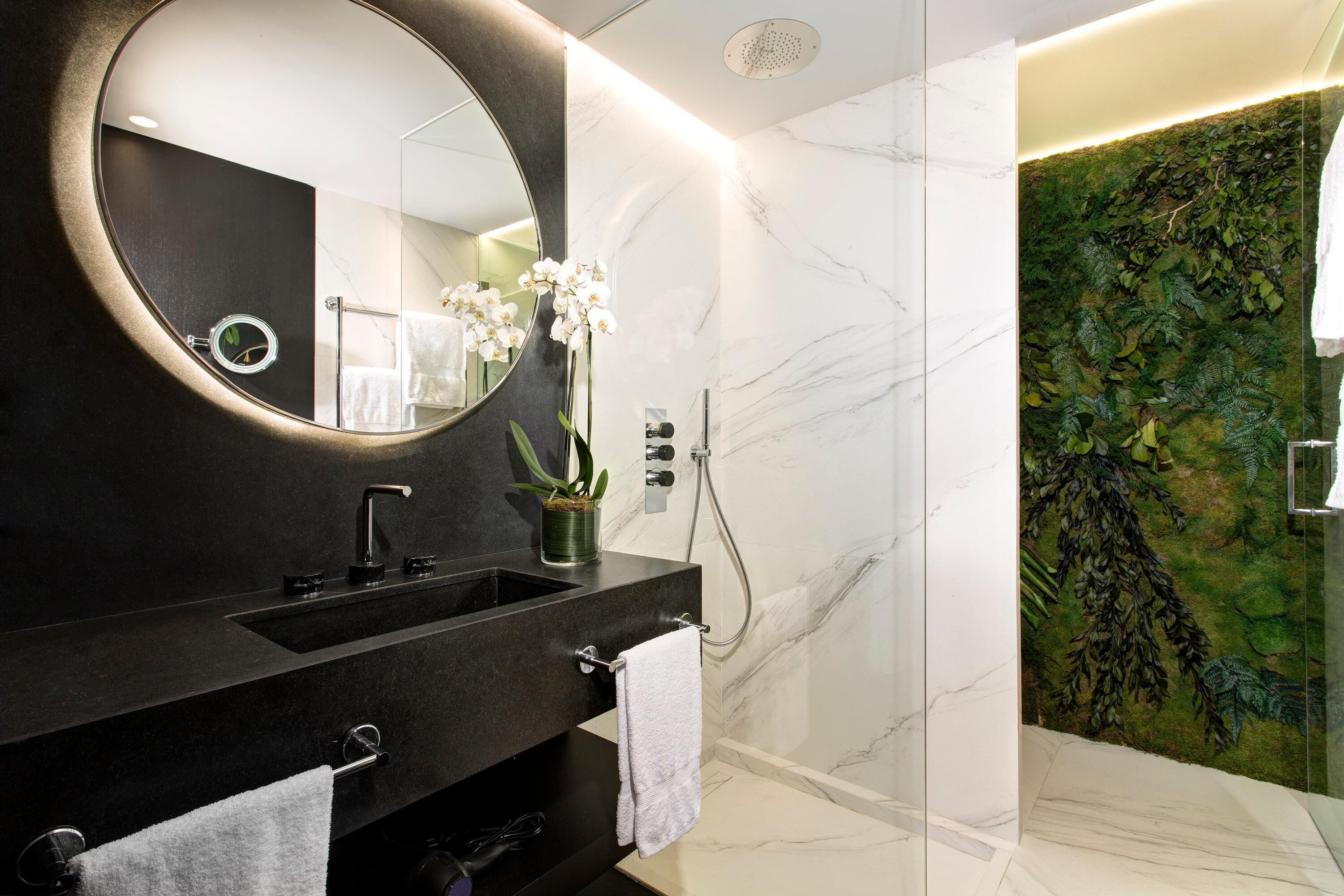 Un baño moderno con tocador negro, un espejo redondo, una orquídea en maceta y una zona de ducha con un diseño de hojas en la pared.