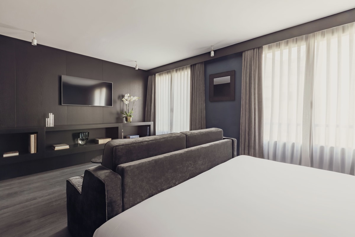 Ein modernes Hotelzimmer mit einem großen Bett, einem Flachbildfernseher, einer Wand aus dunklem Holzpaneel, einer kleinen Sitzecke und einem großen Fenster mit leichtem Vorhang.