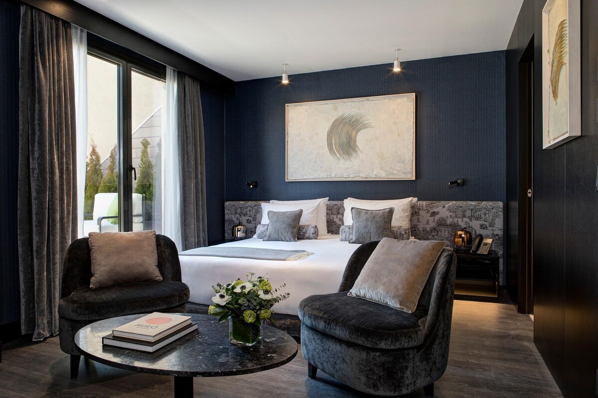 Une chambre moderne avec un grand lit, deux fauteuils, une table basse ronde ornée d’un livre et de fleurs, et une vue sur le paysage urbain par la fenêtre.