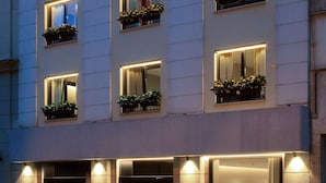 Um hotel moderno com vários andares, cada um com varandas adornadas com plantas em vasos. O edifício tem um design contemporâneo e elegante, com grandes janelas e fachada bem iluminada.