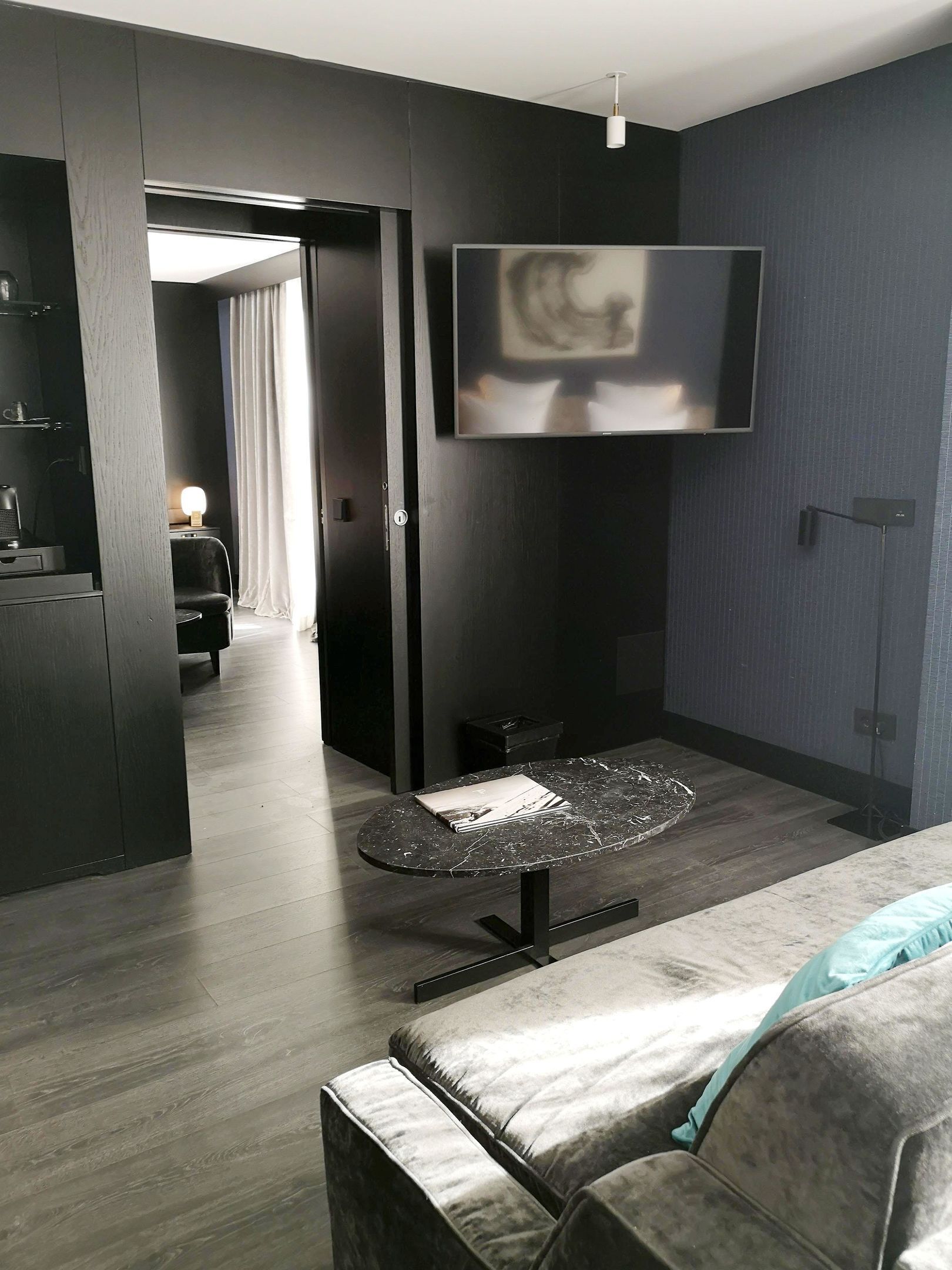 Una habitación de hotel moderna con suelo de madera oscura, un sofá gris, una pequeña mesa redonda y un televisor de pantalla plana montado en la pared.