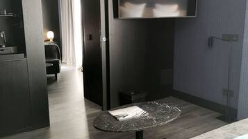 Una habitación de hotel moderna con suelo de madera oscura, un sofá gris, una pequeña mesa redonda y un televisor de pantalla plana montado en la pared.