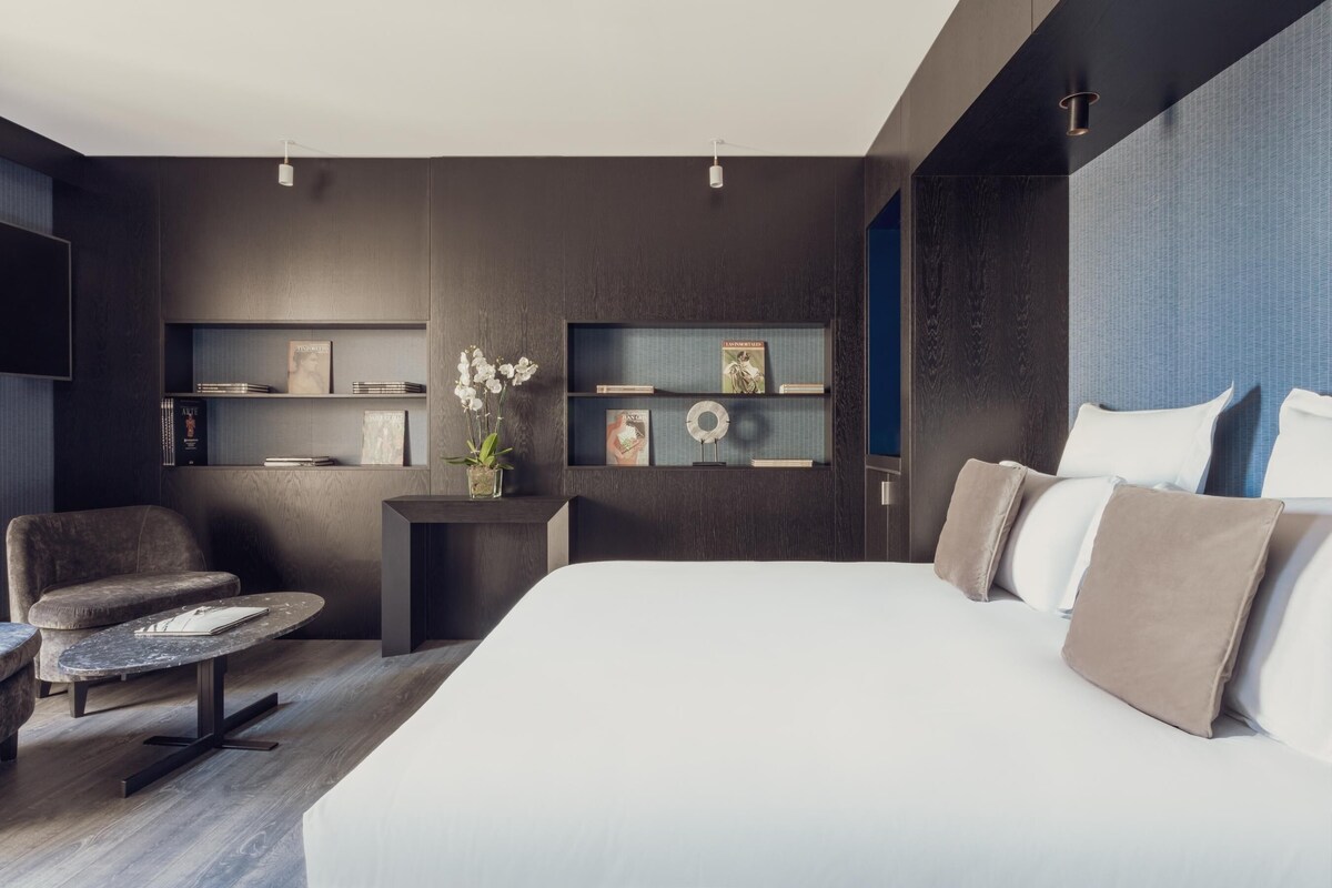 Una habitación de hotel moderna con una cama grande, una zona de estar con una pequeña mesa y estanterías empotradas con libros y objetos decorativos.
