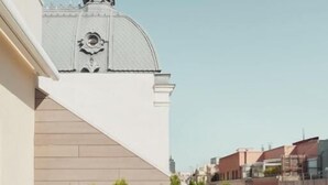 Uma varanda com um sofá branco, uma mesa, uma pequena planta em vaso e vista para um edifício com cúpula.