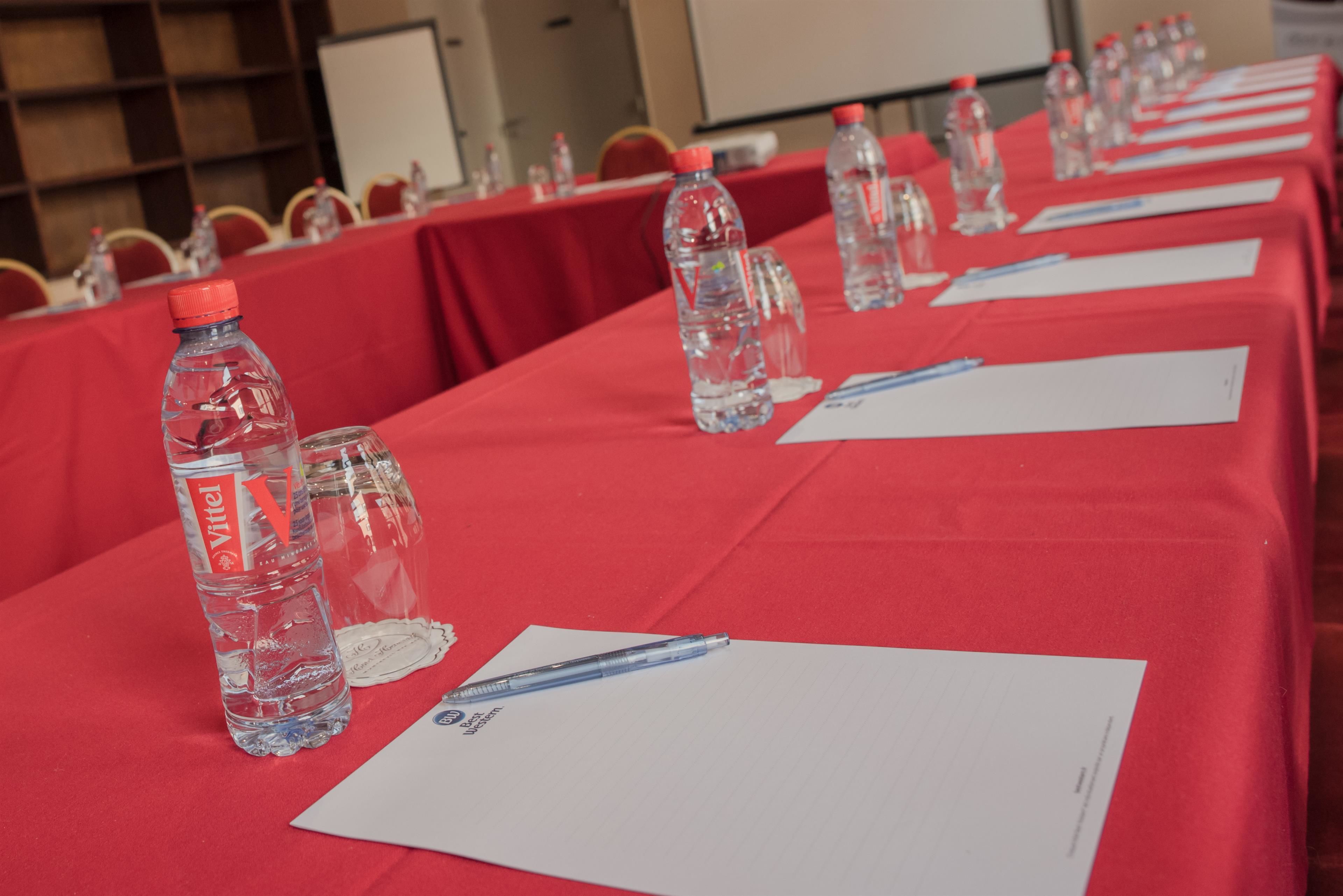 Un salón de conferencias con una larga mesa cubierta con un mantel rojo, botellas de agua y tarjetas de identificación.
