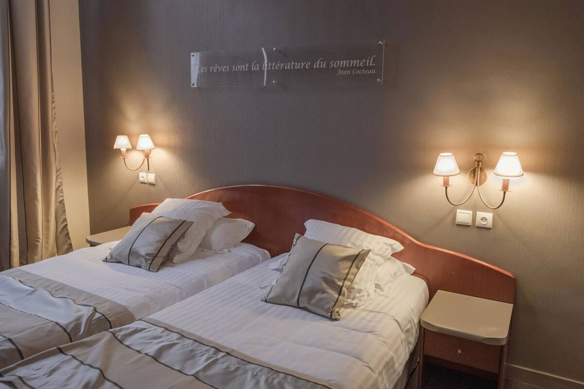 Quarto de hotel com duas camas, uma citação na parede e abajures.