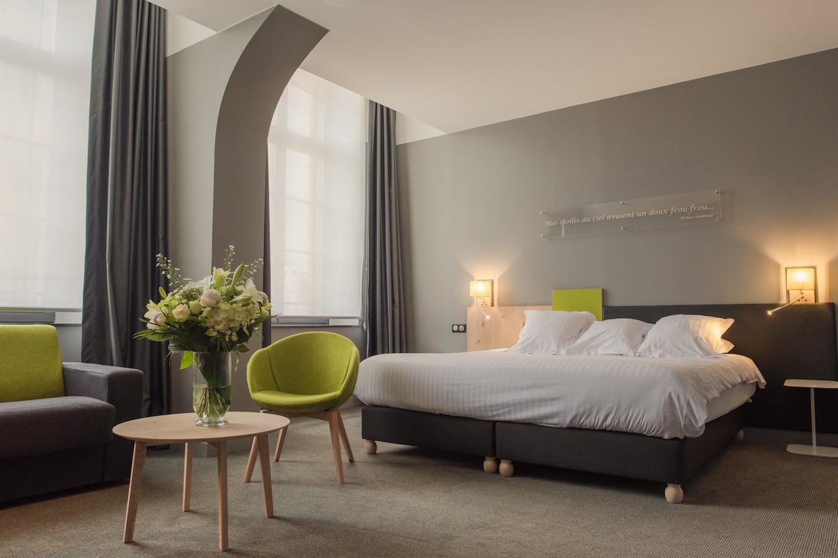Una habitación de hotel moderna con una cama, una silla verde, una mesita con un florero y una mesita de noche con una lámpara.