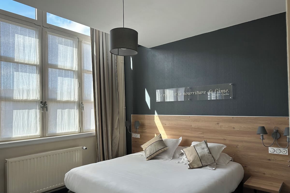 Una habitación de hotel moderna con una cama grande, cabecera de madera, lámpara de pared y ventana con cortinas.