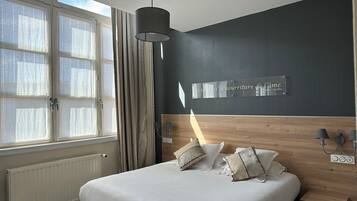 A modern hotel room with a large bed, a wooden headboard, a wall-mounted lamp, and a window with curtains.