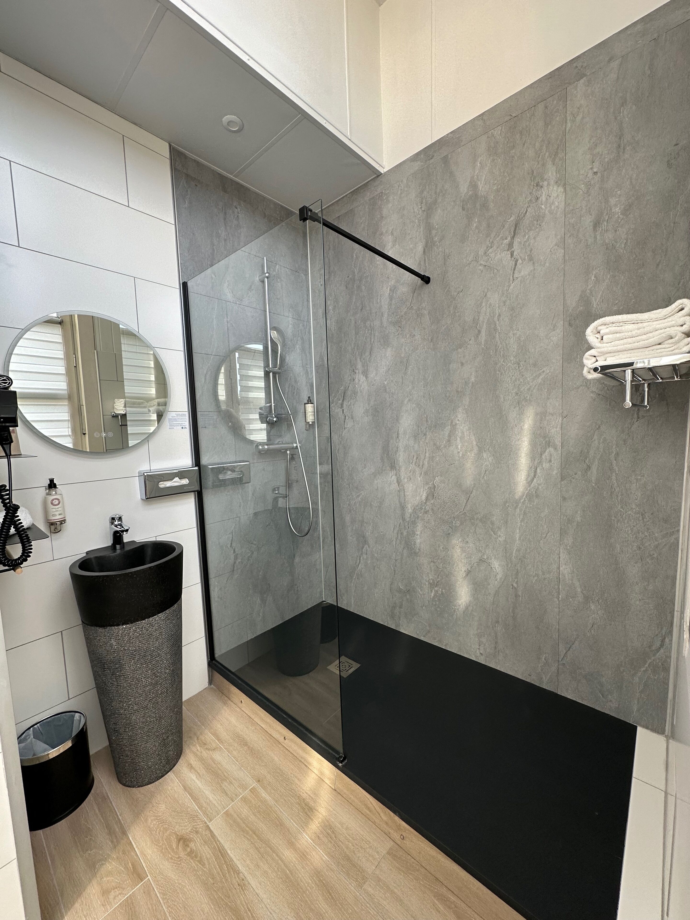 A modern bathroom with a glass-enclosed shower, a round mirror, a towel rack, and a trash bin.