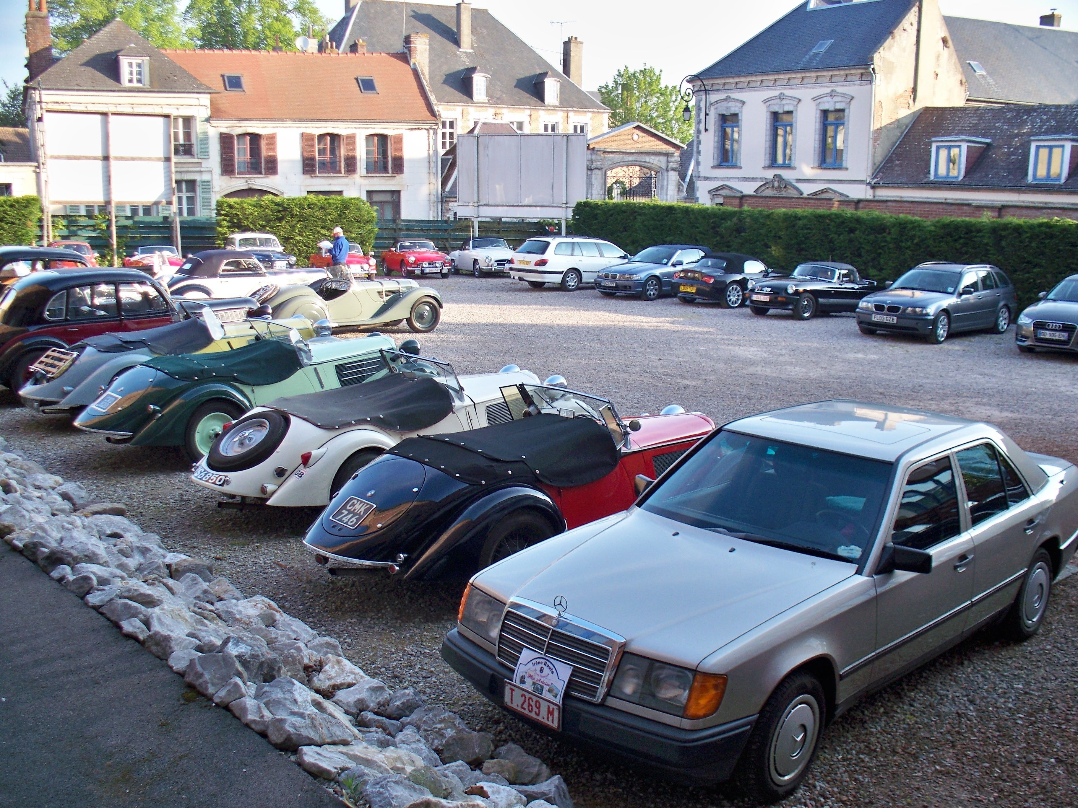 Ein Parkplatz mit verschiedenen Oldtimern und modernen Autos, umgeben von Wohnhäusern.