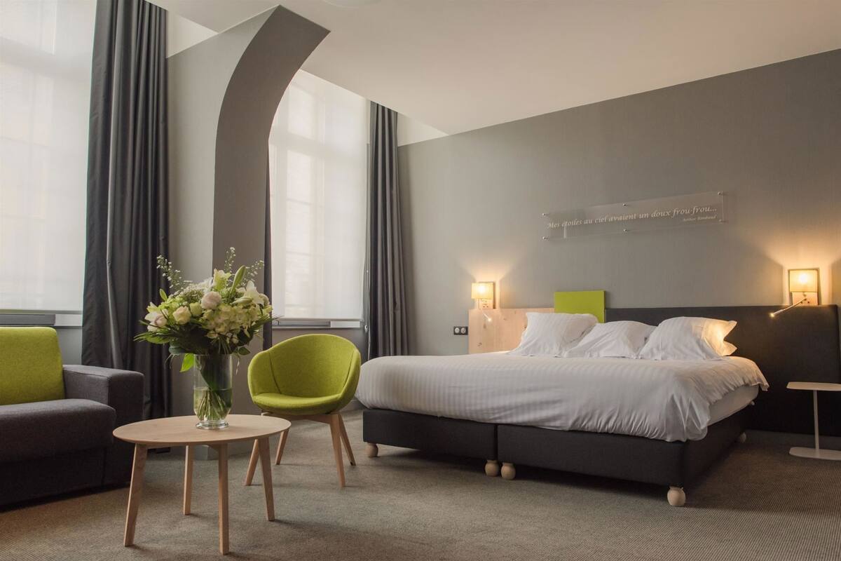 Quarto de hotel moderno com uma cama grande, uma cadeira verde, uma mesinha redonda e um vaso de flores.