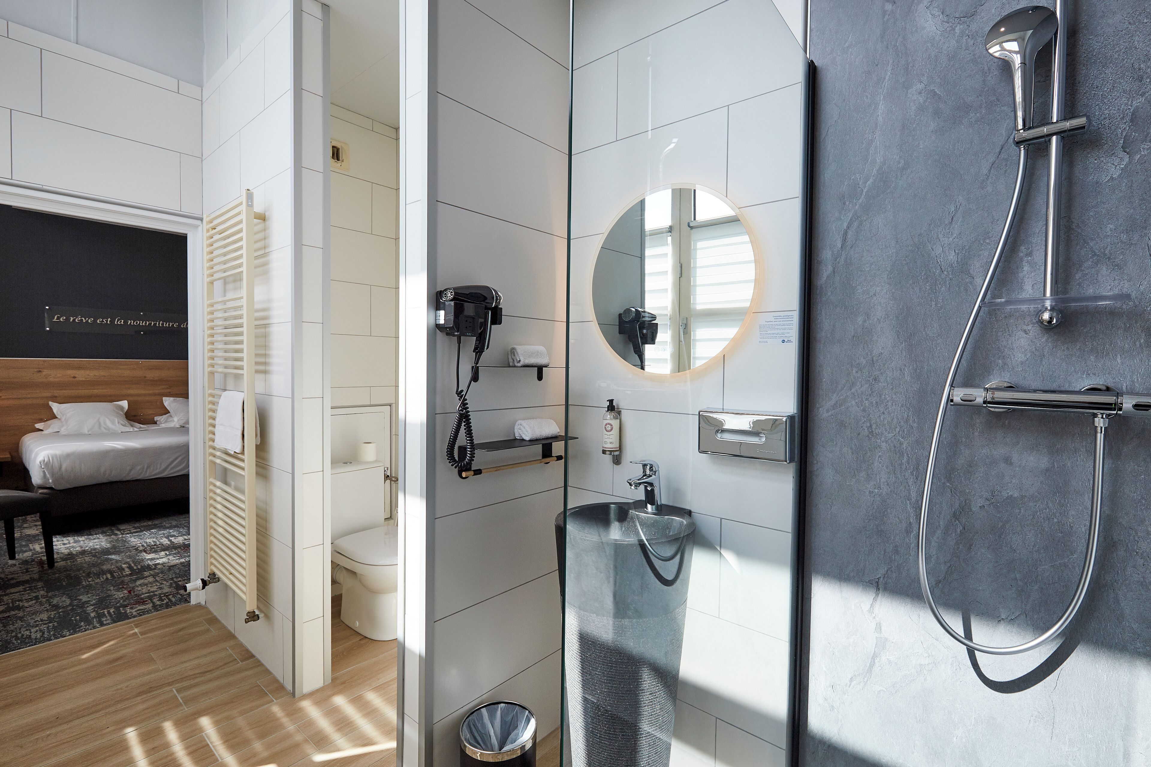 Un baño moderno con ducha de vidrio, un espejo redondo y un secador de pelo en una repisa.