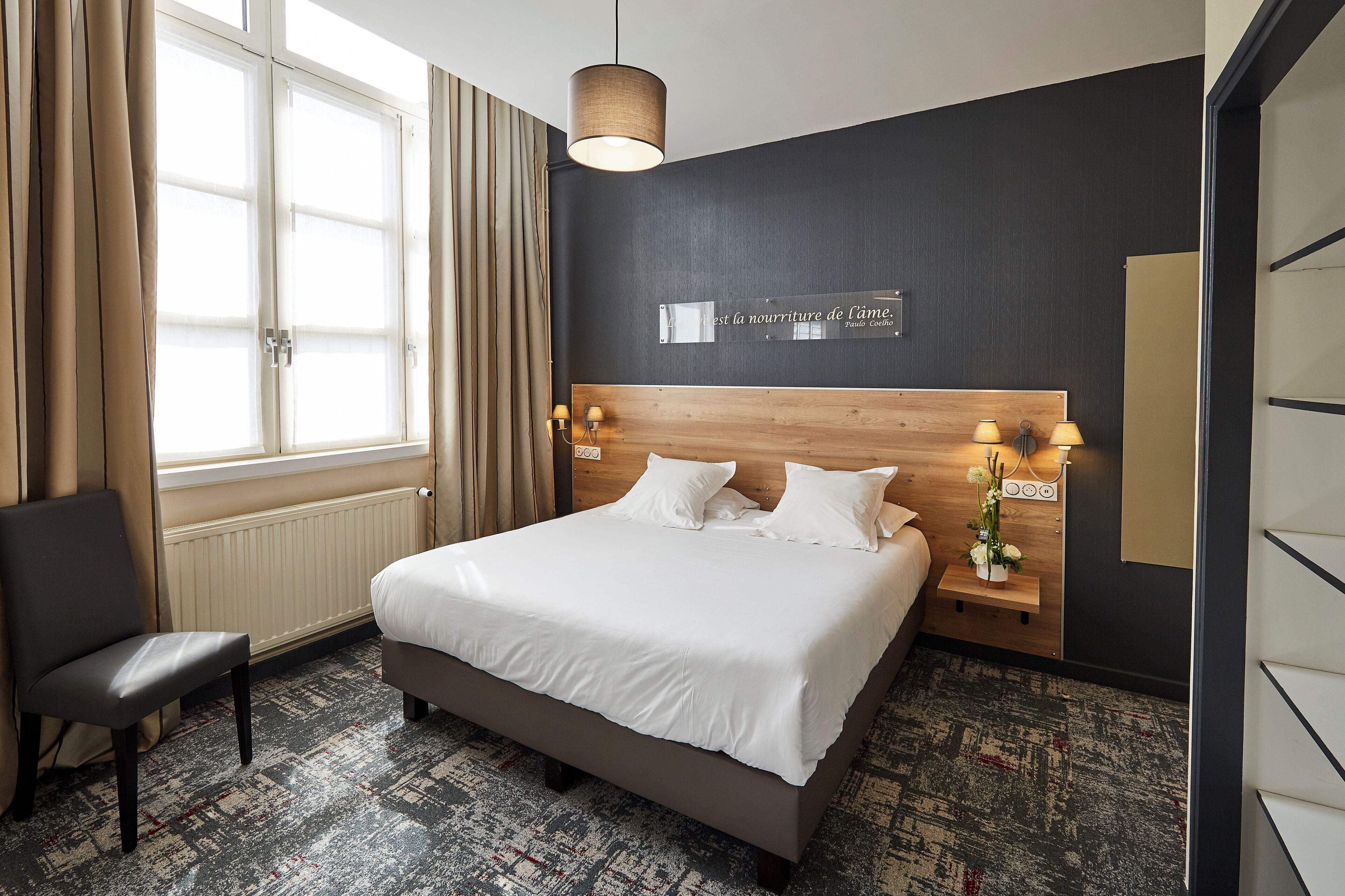 Une chambre d’hôtel avec un grand lit, une tête de lit en bois, une table de chevet avec des lampes, une chaise et une fenêtre avec des rideaux.