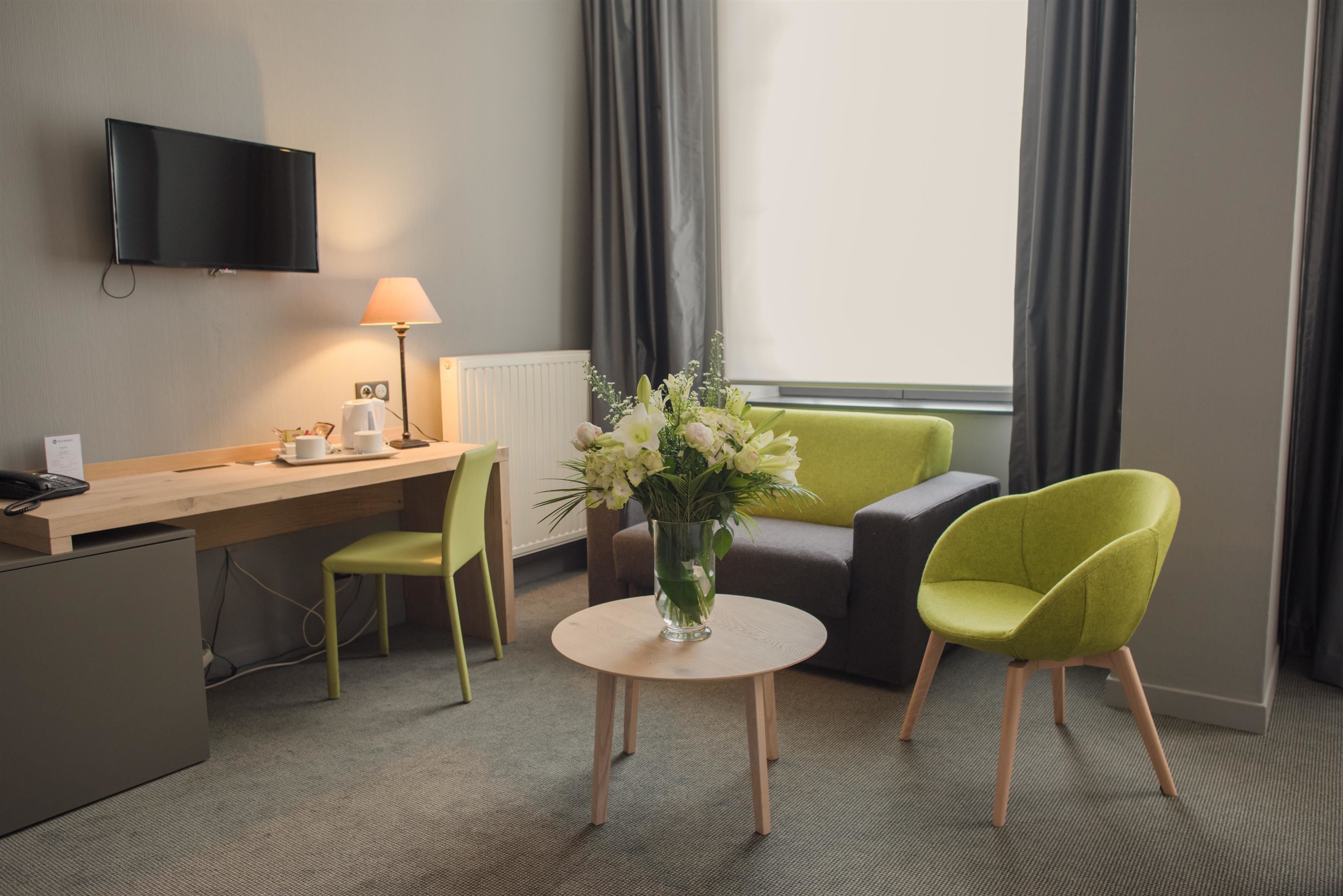 Una habitación de hotel con escritorio, una silla, un sofá, un televisor montado en la pared y una ventana con cortinas.
