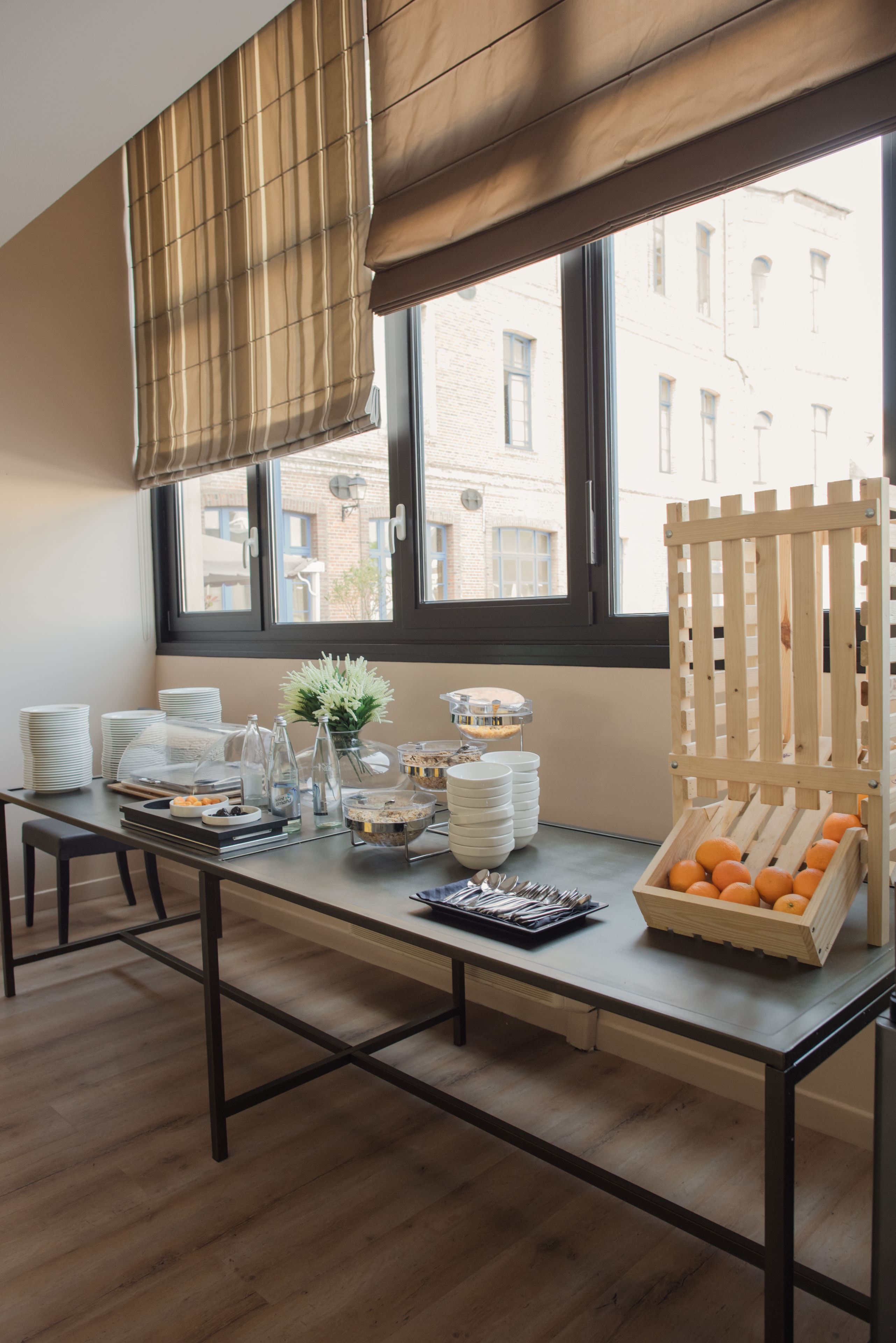 Een ontbijtopstelling met een tafel, borden, kommen en een fruitmandje bij een raam met jaloezieën.