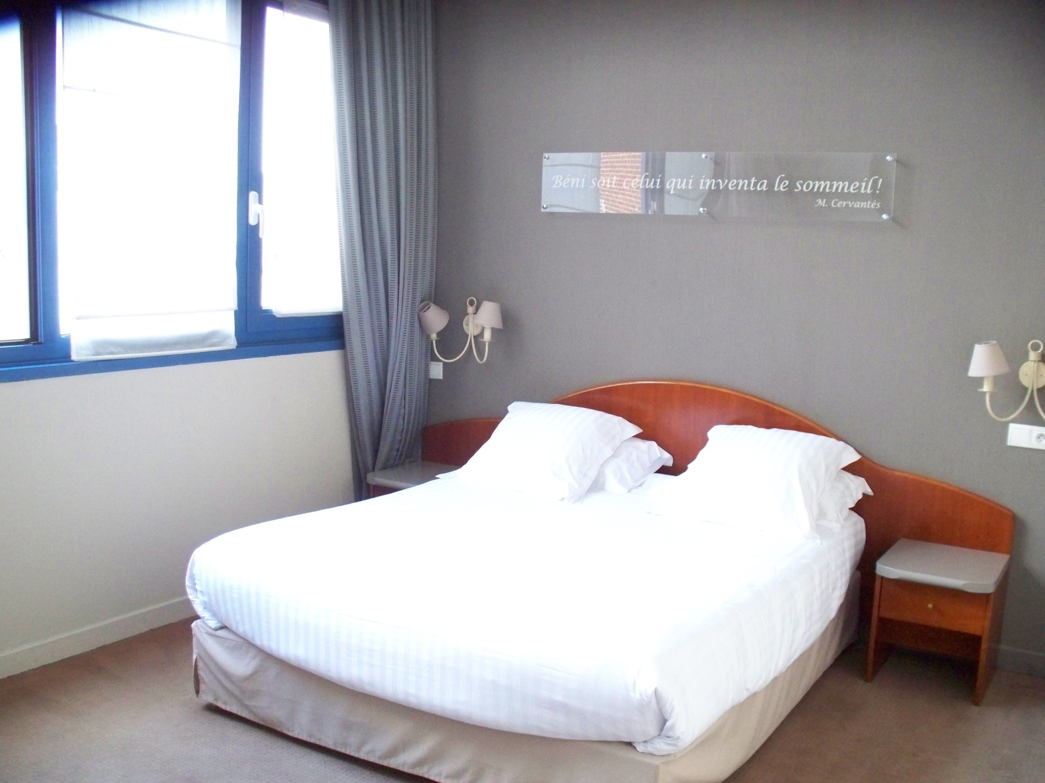 A hotel room with a bed, a nightstand, a lamp, a window with curtains, and a wall sign.