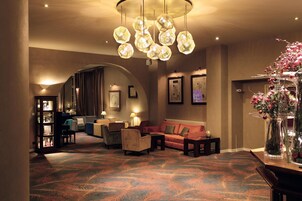 Vestíbulo de hotel con alfombra estampada, un sofá rojo, una mesita de café y un jarrón decorativo con flores.