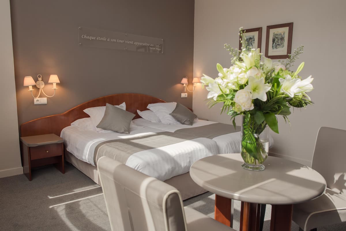 Una habitación de hotel con cama, mesitas de noche, una mesita pequeña con un jarrón de flores y lámparas de pared.