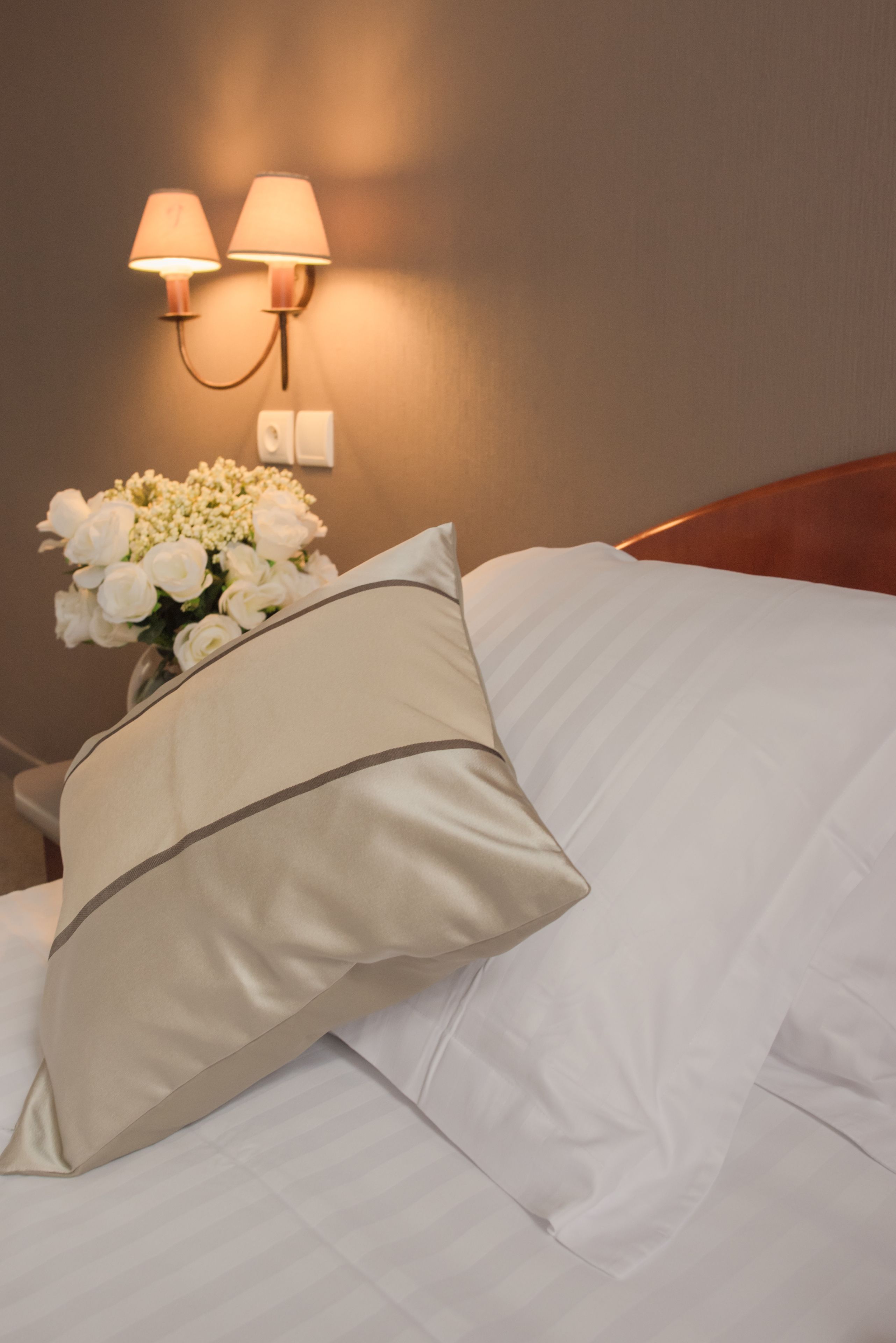A neatly made bed with white linens and a beige pillow, a vase of white flowers, and a wall-mounted lamp.