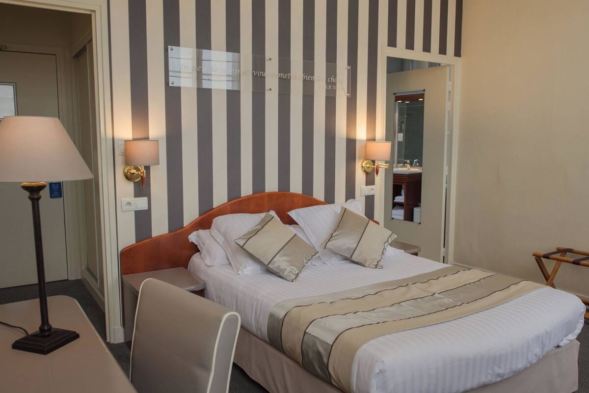 Quarto de hotel com cama, luminárias de cabeceira, escrivaninha e uma cadeira.