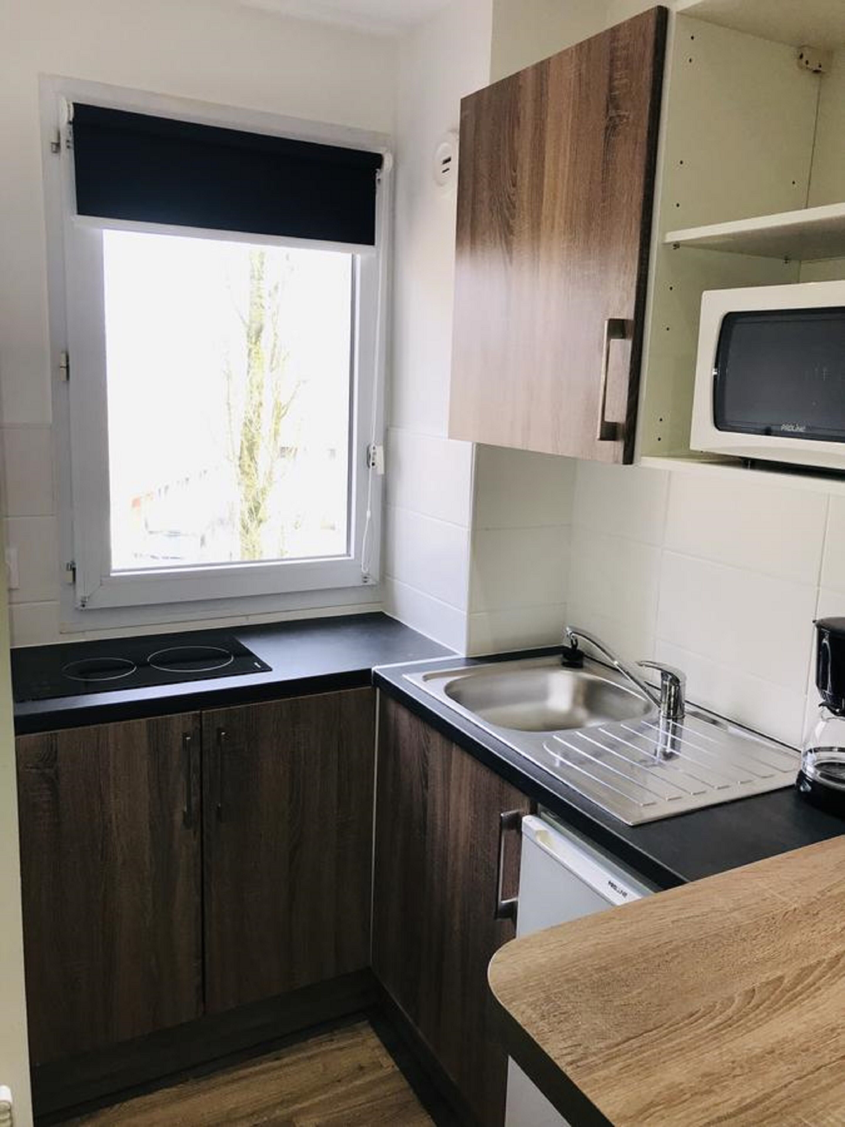 A compact kitchen with dark wooden cabinets, a black countertop, a built-in microwave, and a window with a black roller blind.