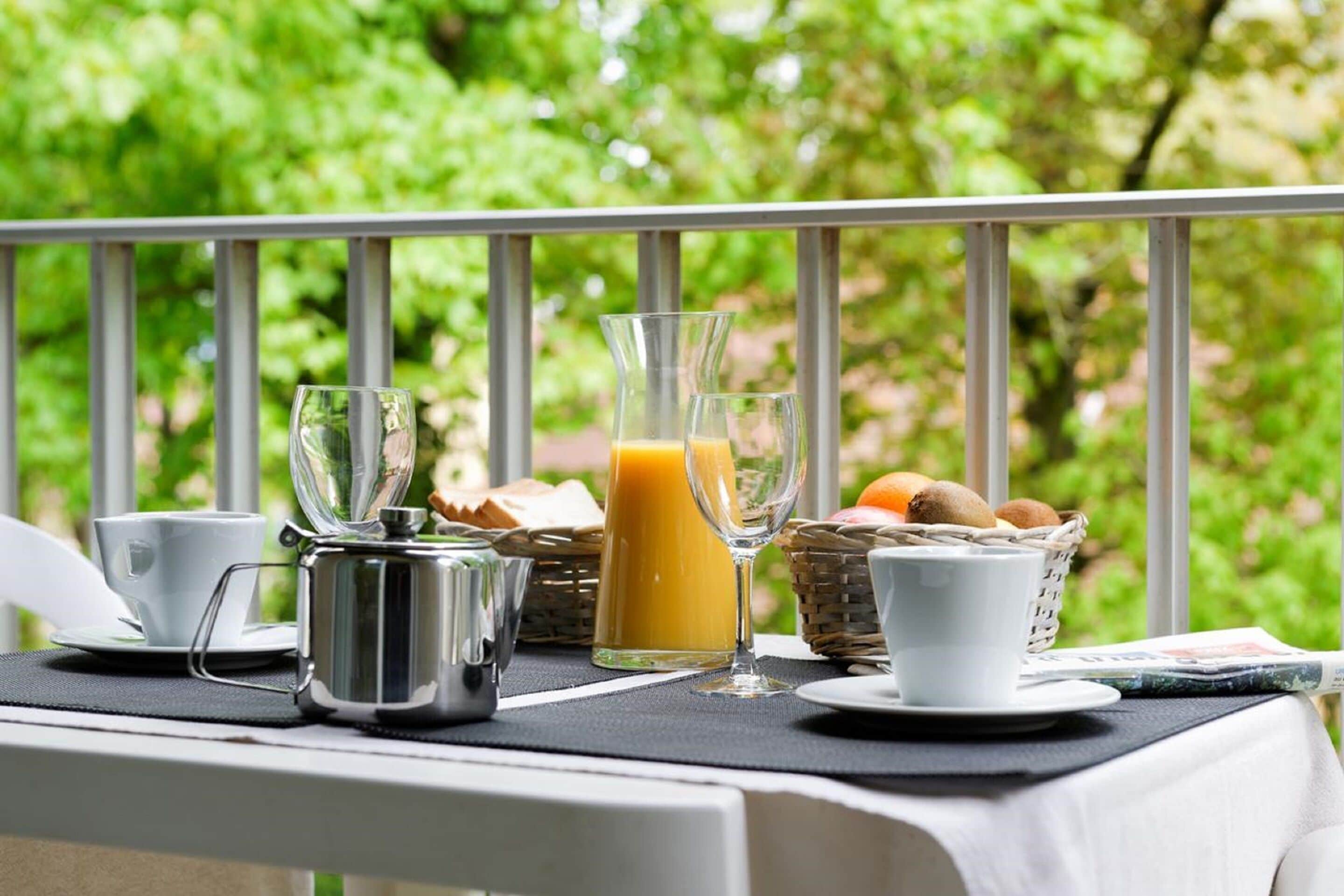 Preparación para el desayuno en un balcón, con una mesa, cafeteras, vasos y una cesta de frutas.
