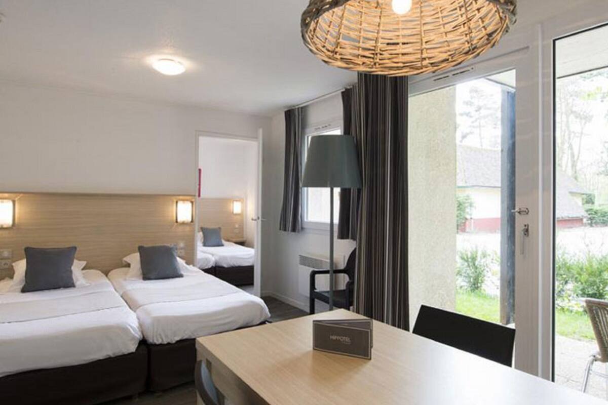 A hotel room with two beds, a dining table, a wicker ceiling light, and a view of the outside.