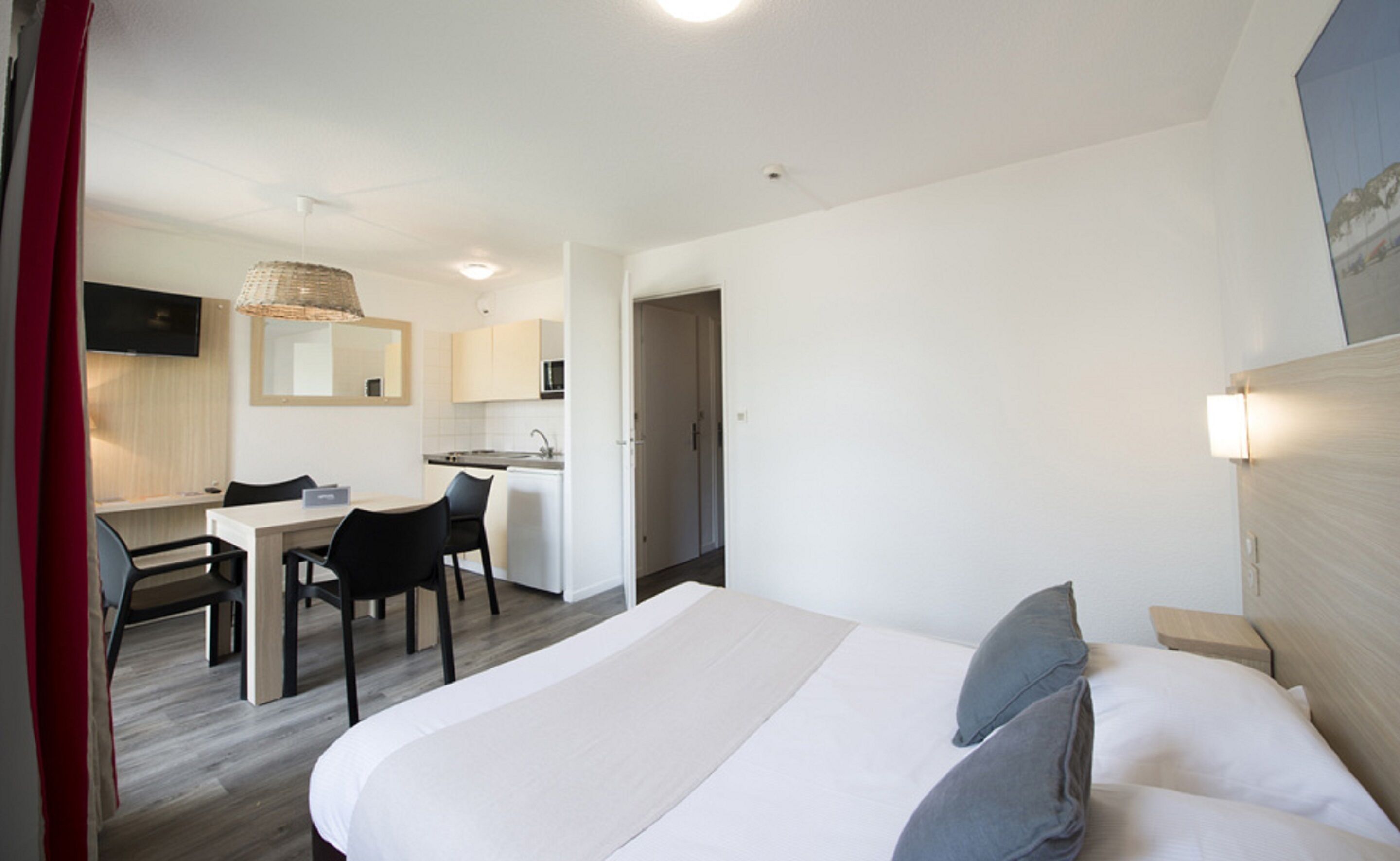 Habitación de hotel con cama, mesita de noche y escritorio con sillas. Se observa una kitchenette al fondo.