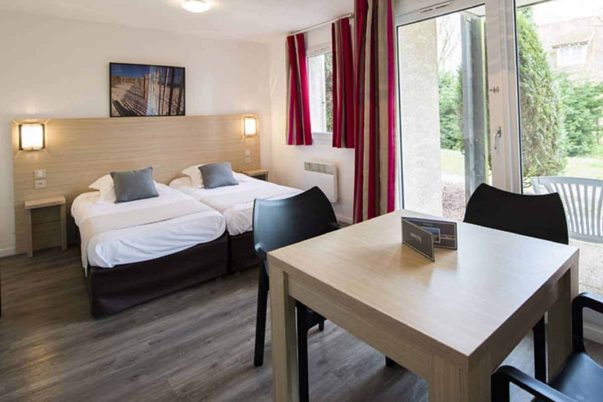 A hotel room with two beds, a table, and a view of the outdoors.